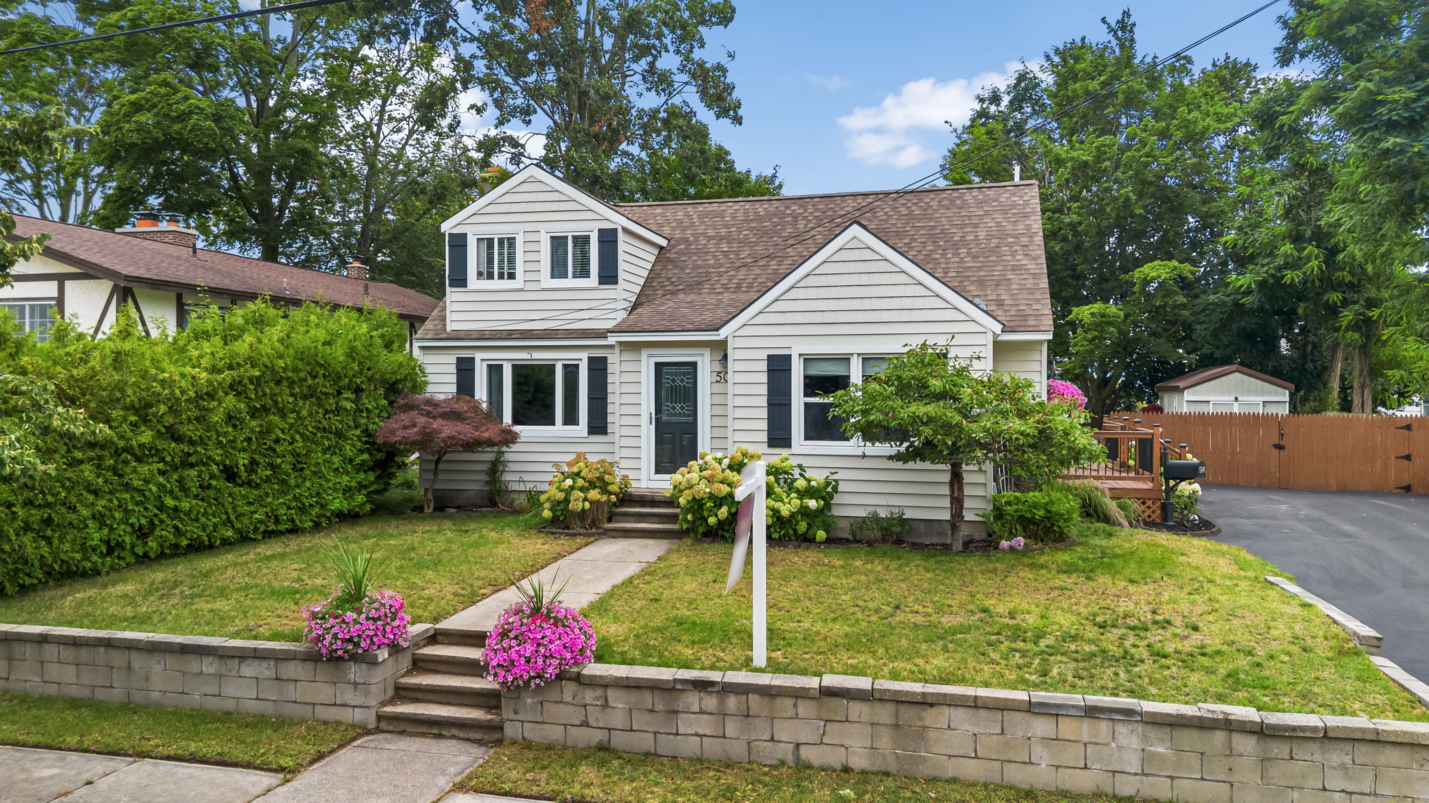 A house with a white exterior, brown roof and a yard with flowers is displayed against a blue sky with trees and a buildin...