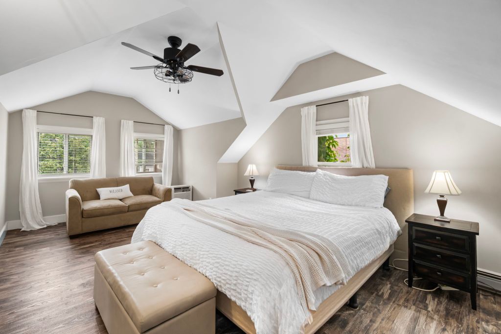 A bedroom featuring a queen-sized bed with a tufted beige leather ottoman and a beige loveseat in the corner.