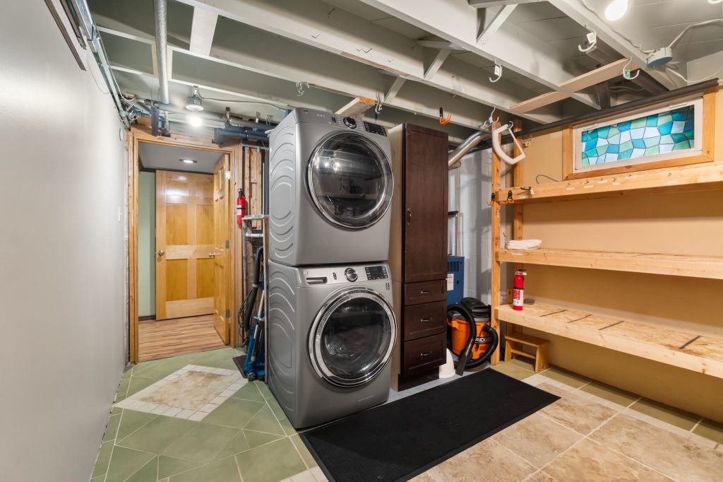 A laundry room featuring a washer and dryer stacked on top of each other, with a black mat in front, surrounded by a bench...
